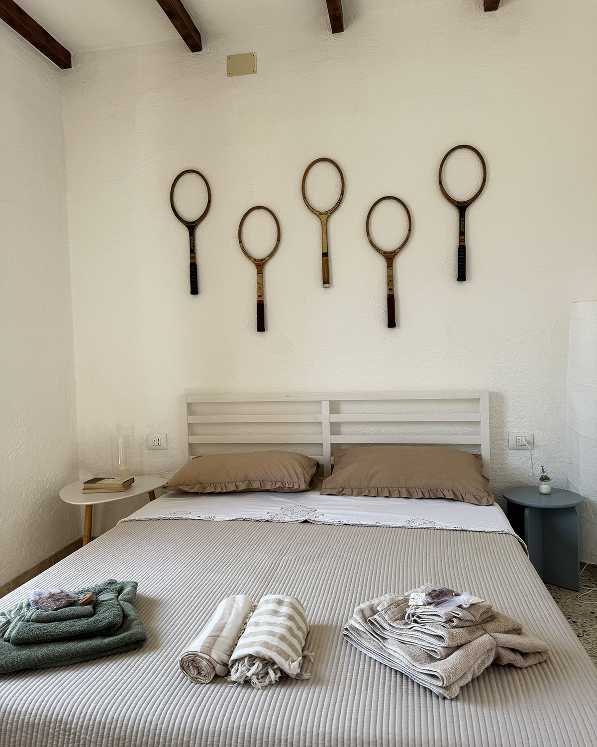 Camera matrimoniale di Mae a Favignana con letto bianco, biancheria dai toni neutri e racchette vintage decorative alla parete.