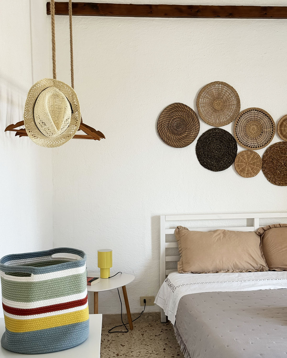 Dettaglio di una camera di Mae a Favignana con letto bianco, cuscini beige, cappello in paglia e decorazioni intrecciate alla parete.