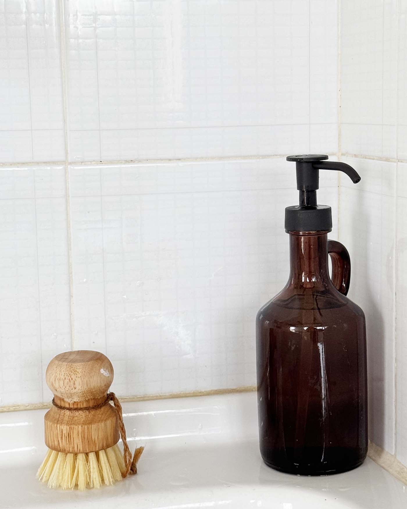 Dettaglio del lavello di Mae a Favignana con dispenser in vetro ambrato e spazzola in legno su piastrelle chiare.