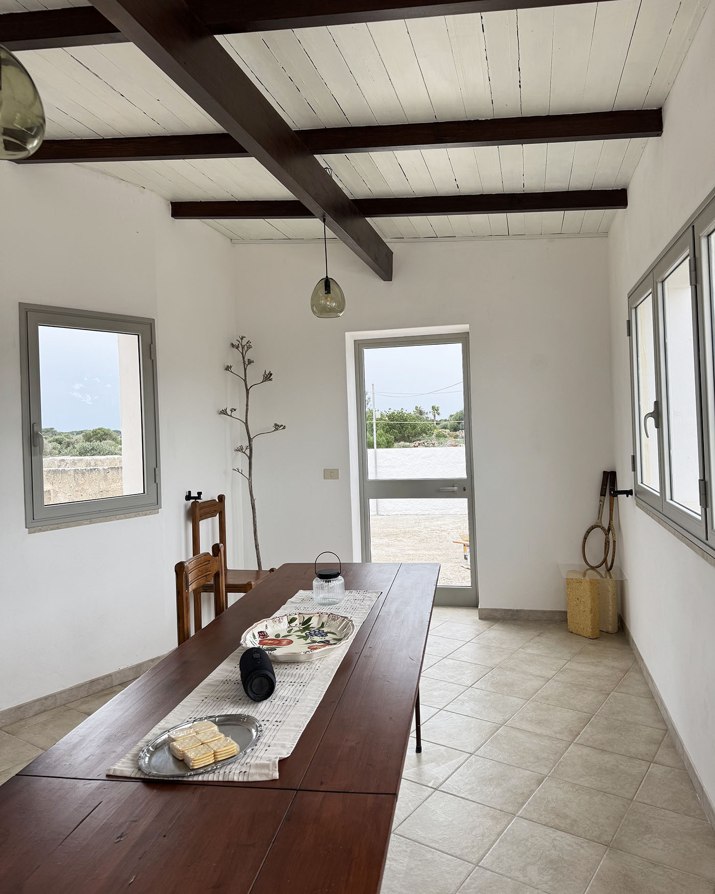 Spazio pranzo interno di Mae a Favignana con lungo tavolo in legno, travi a vista e accesso diretto verso l’esterno.